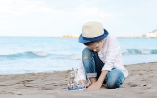 Картинка Маленький мальчик в шляпе играет с игрушечным корабликом на песке
